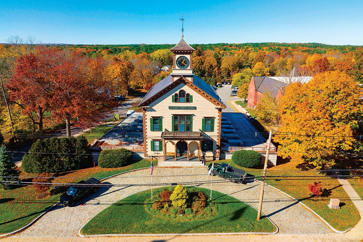 A historic two-story town hall building with a clock tower and a weather vane on top, labeled "Acton Town Hall." The building features beige siding with brown trim and green shutters. It is surrounded by a circular driveway with a small landscaped island in the center, displaying an American flag and another flag. The scene is set in autumn with colorful fall foliage in shades of orange, yellow, and red. Two vehicles are parked near the building, and the sky is clear and blue.