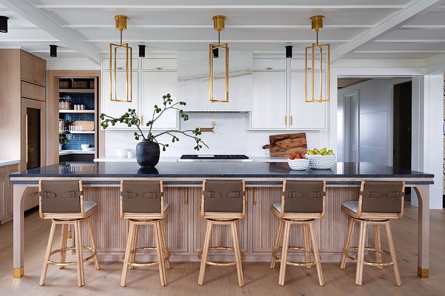 Modern kitchen with a large island featuring a dark countertop and light wood paneling. Five wooden bar stools with brown leather backs are arranged along the island. Above the island, three gold-framed pendant lights hang from a white coffered ceiling. The back wall has white cabinetry with gold handles, a marble range hood, and a built-in stovetop. A black vase with green branches and two white bowls with fruit are placed on the island. Light wood flooring completes the space.