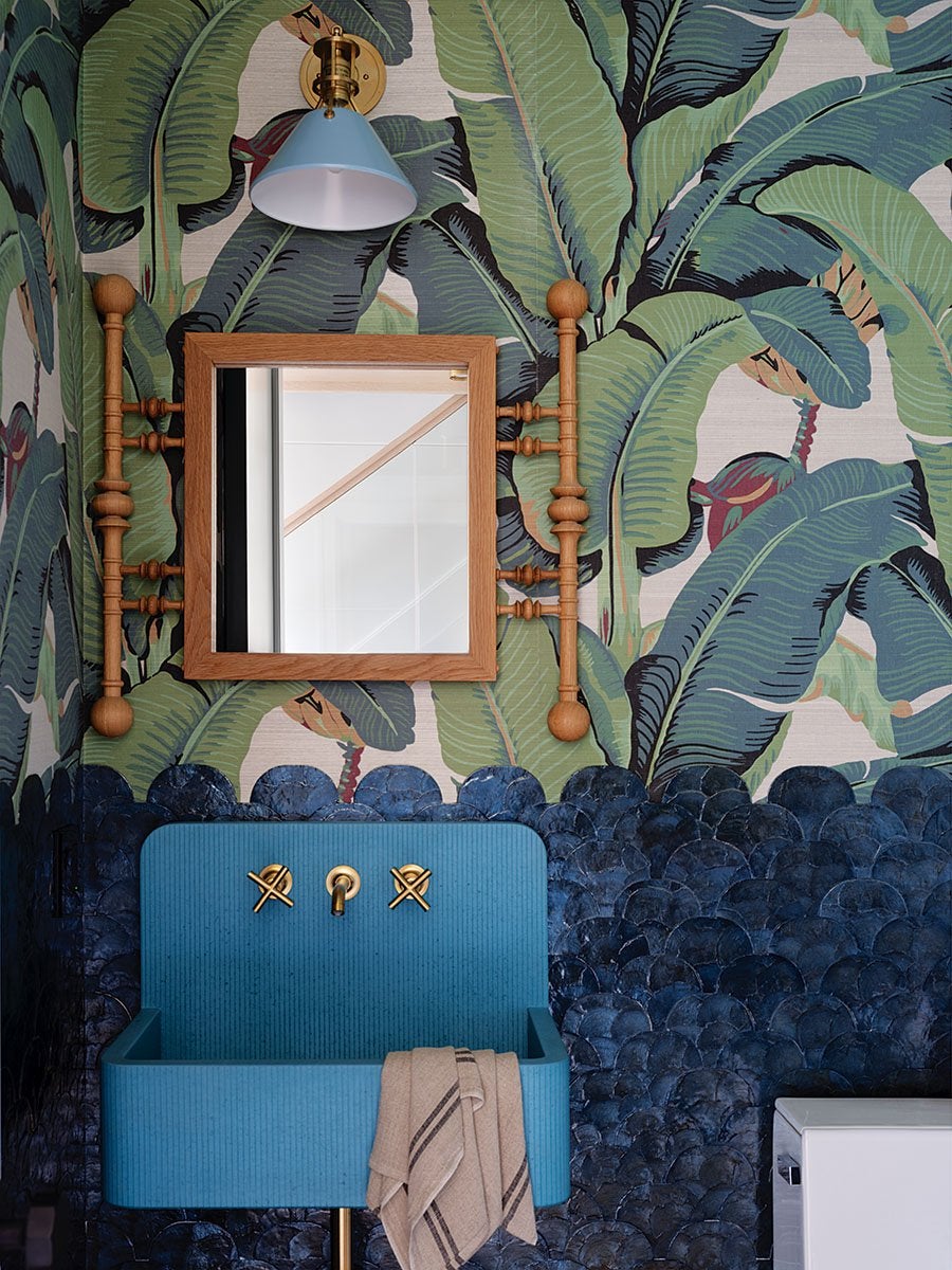 A bathroom corner featuring a blue ribbed sink with gold faucet handles and a beige towel draped over the edge. Above the sink is a wooden-framed square mirror with turned wooden side supports. The wall behind has a tropical leaf wallpaper in shades of green and blue, with a textured dark blue scalloped tile halfway up. A brass and blue wall sconce light fixture is mounted above the mirror.