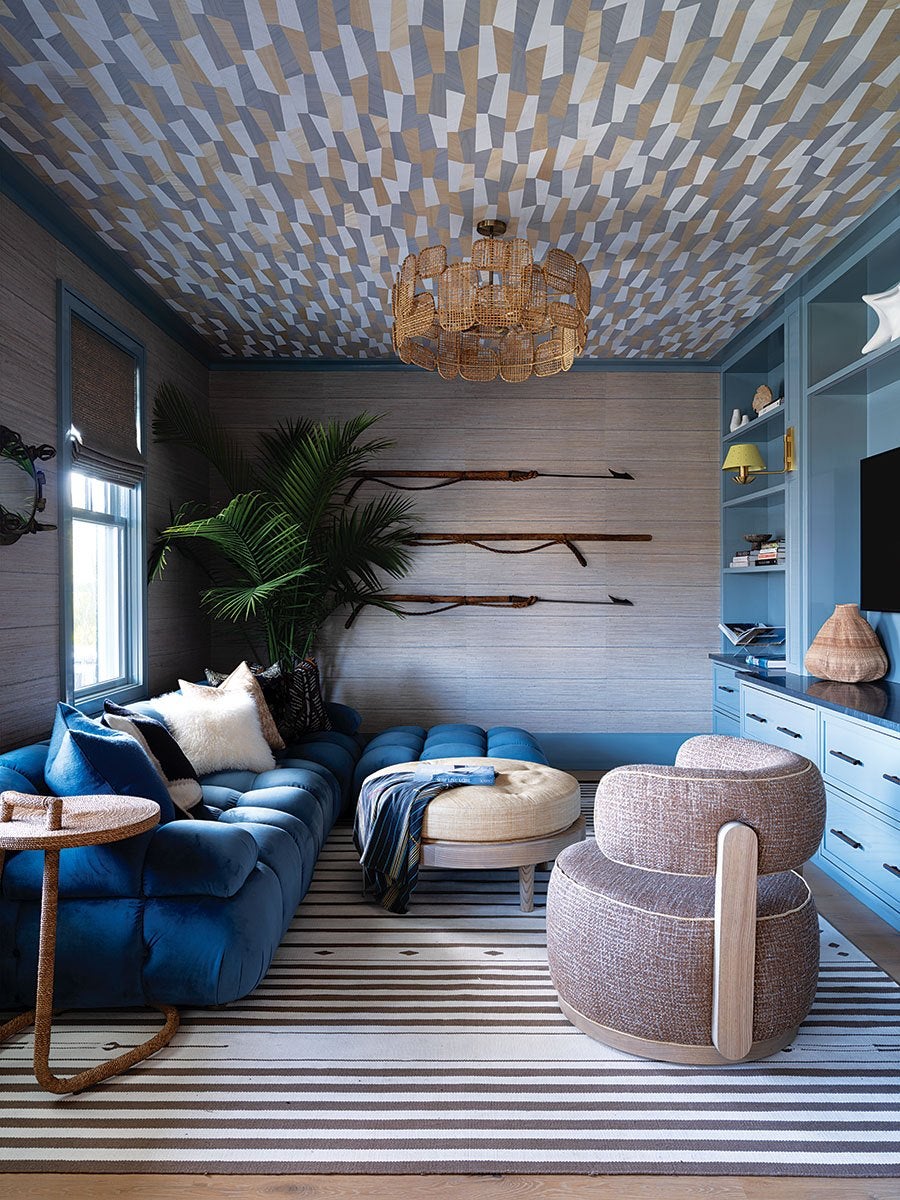 Living room with a blue tufted sectional sofa adorned with various pillows, a round beige ottoman with a blue throw, and a textured round armchair with wooden accents. The room features a striped area rug, a geometric patterned ceiling, and a woven chandelier. The walls are covered in light wood paneling, with three decorative wooden spears mounted on one wall. Built-in blue cabinetry with shelves and drawers is on the right side, and a large green plant is positioned near a window with a blue frame and a woven shade.