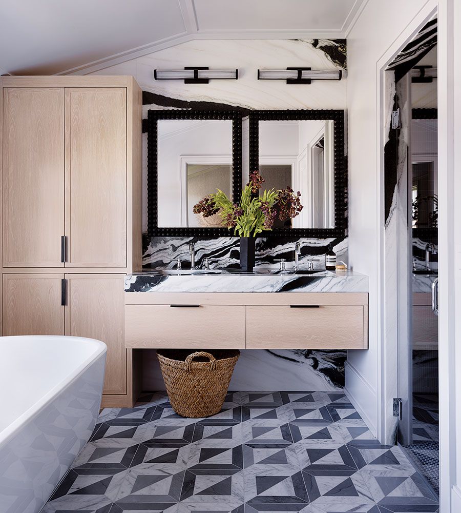 Bathroom vanity with light wood cabinets and drawers, a white marble countertop with black veining, and two black-framed mirrors above. A black vase with green and dark red flowers is centered between two chrome faucets. The backsplash features a striking black and white marble pattern. The floor has a geometric tile design in shades of gray and white. A woven basket is placed under the floating vanity. To the left, part of a white freestanding bathtub is visible. To the right, a doorway leads to a shower area with a glass door.