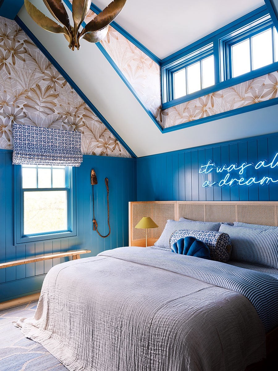 Bedroom with blue paneled walls and floral wallpaper on the slanted ceiling. The bed has a light beige quilt, blue and white striped bedding, and decorative pillows in blue patterns. A wooden headboard with a woven cane design is behind the bed. A neon sign on the wall reads "it was all a dream." A window with a blue and white patterned Roman shade is on the left wall. A wooden bench is beneath the window, and a gold-colored lamp with a yellow shade is on a nightstand next to the bed. The ceiling light fixture has a leaf-like design in a metallic finish.