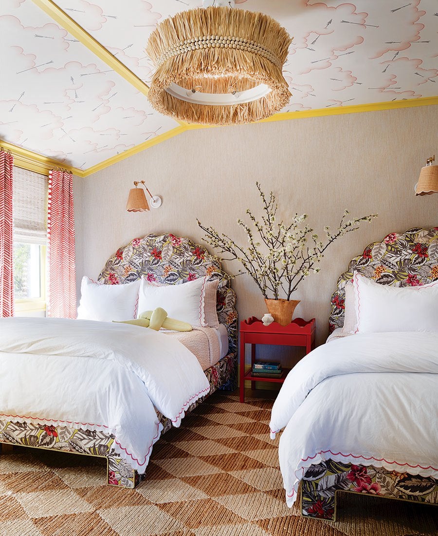 A cozy bedroom features two twin beds with floral-patterned headboards and matching bed frames. Both beds are dressed in white bedding with scalloped pink edges. Between the beds is a red nightstand holding a terracotta vase filled with blossoming branches. The room has beige walls, a ceiling with a whimsical cloud and arrow pattern, and yellow trim. A woven, fringed light fixture hangs from the ceiling. The floor is covered with a textured, diamond-patterned rug in natural tones. Red and white chevron curtains frame a window on the left side.