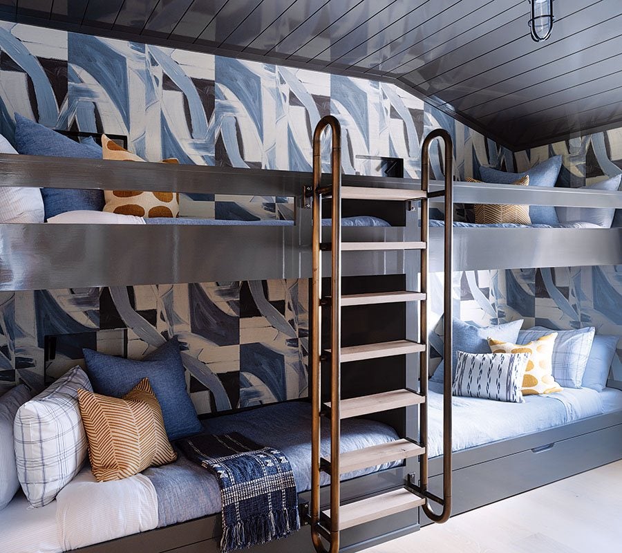 Modern bunk beds with a dark gray frame and wooden ladder, featuring blue and white bedding and a variety of patterned pillows. The wall behind has an abstract blue, white, and black design, and the ceiling is dark gray with a hanging light fixture.