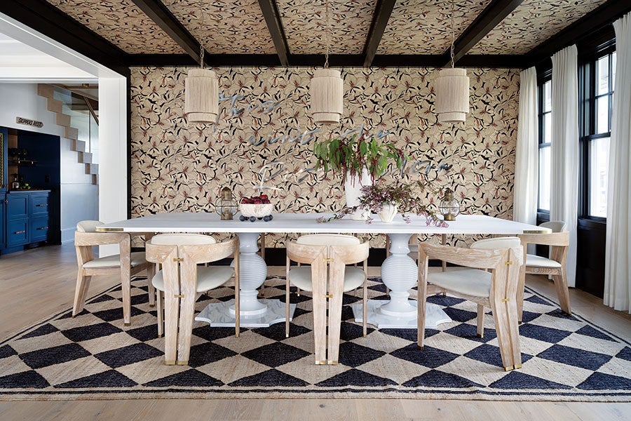 Dining room with a white rectangular table supported by two ornate pedestal bases, surrounded by six light wood chairs with white cushions. The floor features a large black and beige diamond-patterned rug. The back wall and ceiling are covered in floral wallpaper with beige, black, and red tones. Three cylindrical pendant lights hang above the table. Large windows with white curtains are on the right side, allowing natural light into the room. A vase with green and purple flowers is placed on the table.