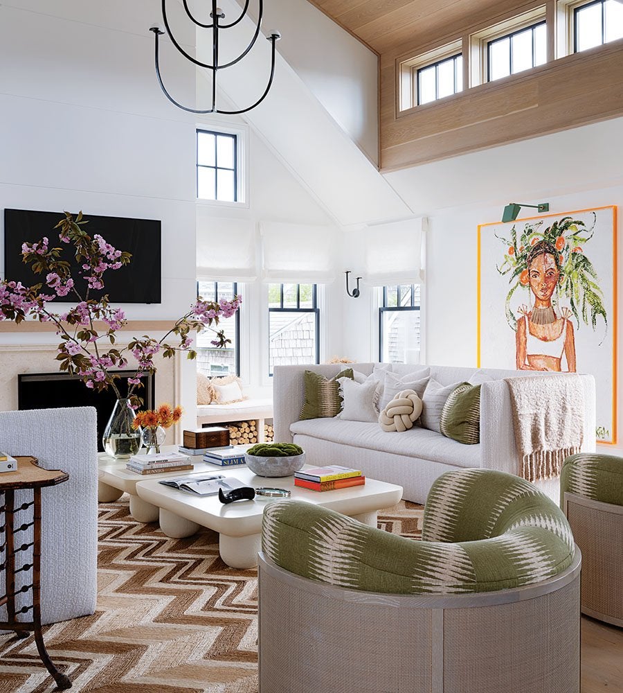 A bright living room with a white sofa adorned with multiple pillows and a throw blanket. In front of the sofa is a low, white coffee table with books, a bowl, and decorative items. Two green patterned armchairs are positioned nearby. The room features a large zigzag-patterned rug in shades of brown and beige. A vase with pink flowers and a smaller vase with orange flowers sit on a side table. A fireplace with a mounted TV above it is on one wall, and a colorful portrait painting with an orange frame hangs on another. The space has large windows with white Roman shades and a high ceiling with wood paneling and additional windows.