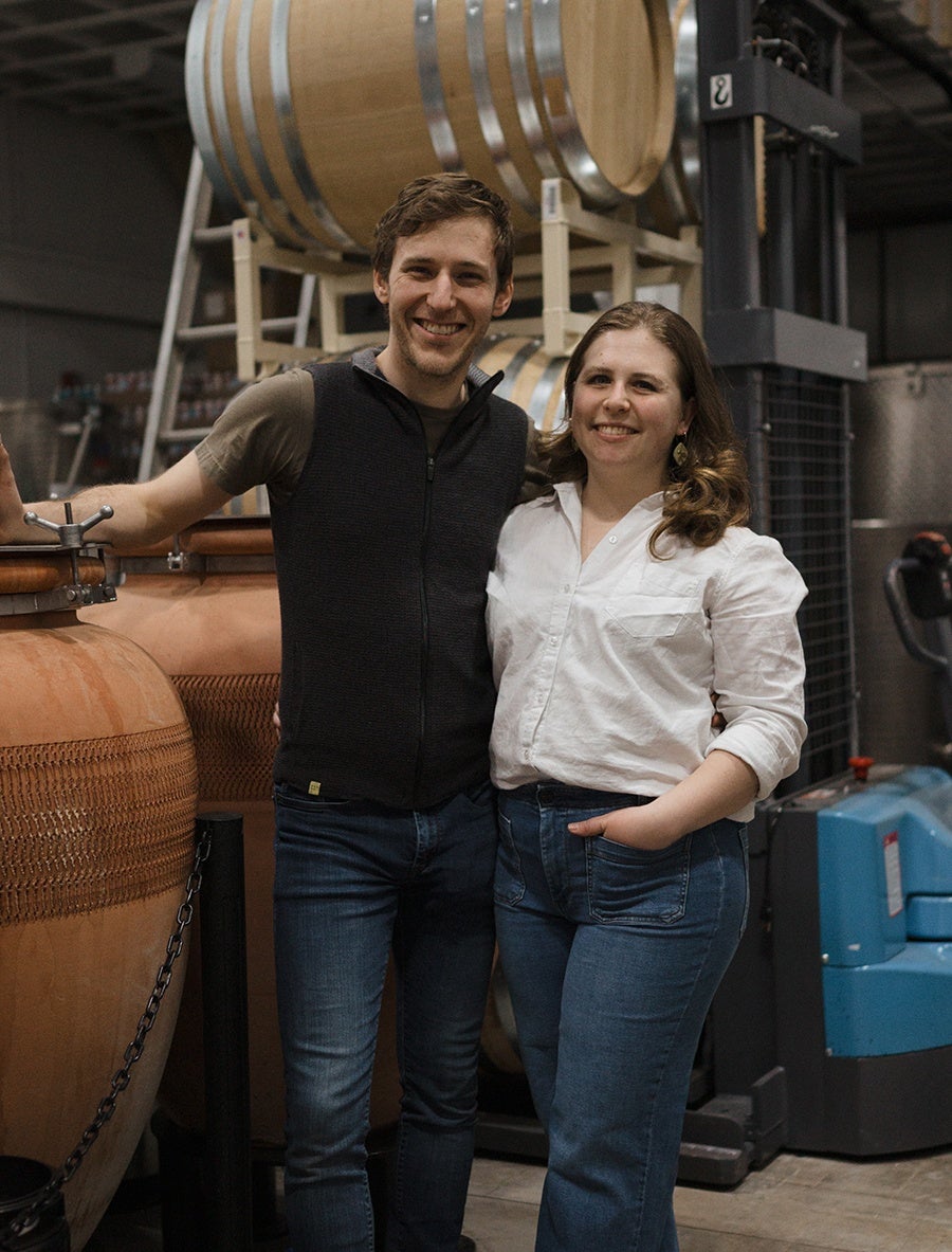 A smiling man and woman standing close together in a winery or brewery setting. The man is wearing a black vest over a brown shirt and blue jeans, while the woman is dressed in a white button-up shirt with rolled-up sleeves and blue jeans. Behind them are large wooden barrels and industrial equipment.