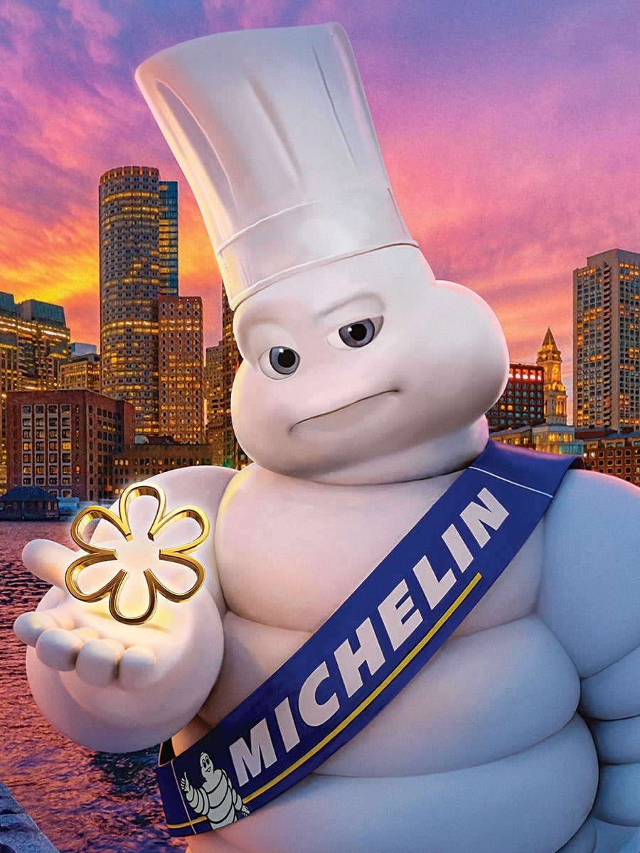 The Michelin Man character wearing a tall white chef's hat and a blue sash with the word "MICHELIN" in white letters, holding a glowing golden Michelin star symbol in his hand. The background features a city skyline at sunset with colorful purple and orange skies.