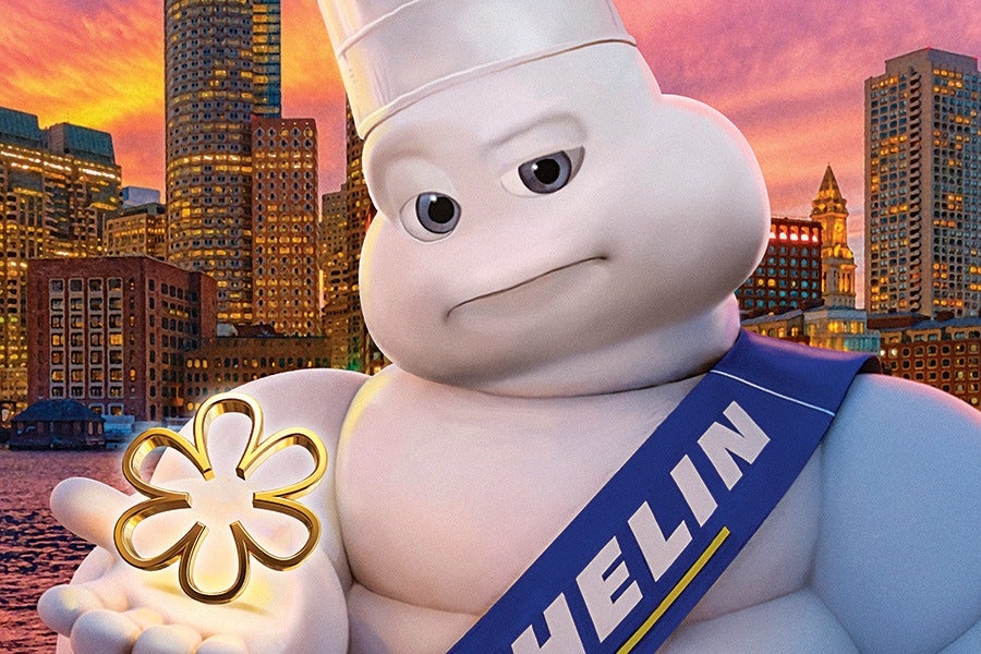 The Michelin Man mascot wearing a white chef's hat and a blue sash with the word "MICHELIN" in white letters, holding a golden flower-shaped object in his hand, with a city skyline and a colorful sunset in the background.