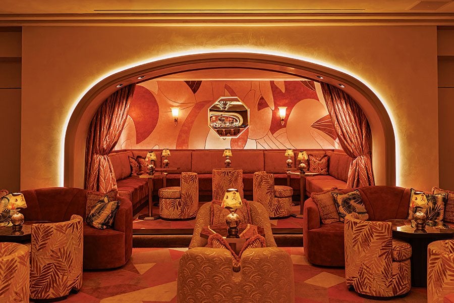 A warmly lit lounge area with an arched entrance framed by soft glowing lights. Inside, there is a U-shaped red velvet sofa along the back wall, accompanied by small round tables and patterned stools. The walls feature abstract, warm-toned artwork and two wall sconces. The seating area in the foreground includes plush armchairs and chairs with leaf-patterned upholstery, each paired with small tables topped with decorative lamps. The overall ambiance is cozy and inviting with rich red and orange hues.