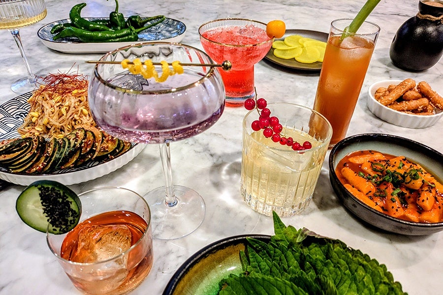 A marble table set with various dishes and cocktails. There are four cocktails: a purple drink in a coupe glass with a lemon twist garnish, a red drink in a short glass with a small orange garnish, a light yellow drink in a rocks glass with red berries garnish, and an amber drink in a tall glass with a green stalk garnish. Food includes a bowl of orange sauce with rice cakes topped with green onions, a plate of grilled zucchini slices and bean sprouts, a dish of green chili peppers, a small bowl of yellow pickled radish slices, a bowl of pretzel sticks, and a plate with fresh green leaves.