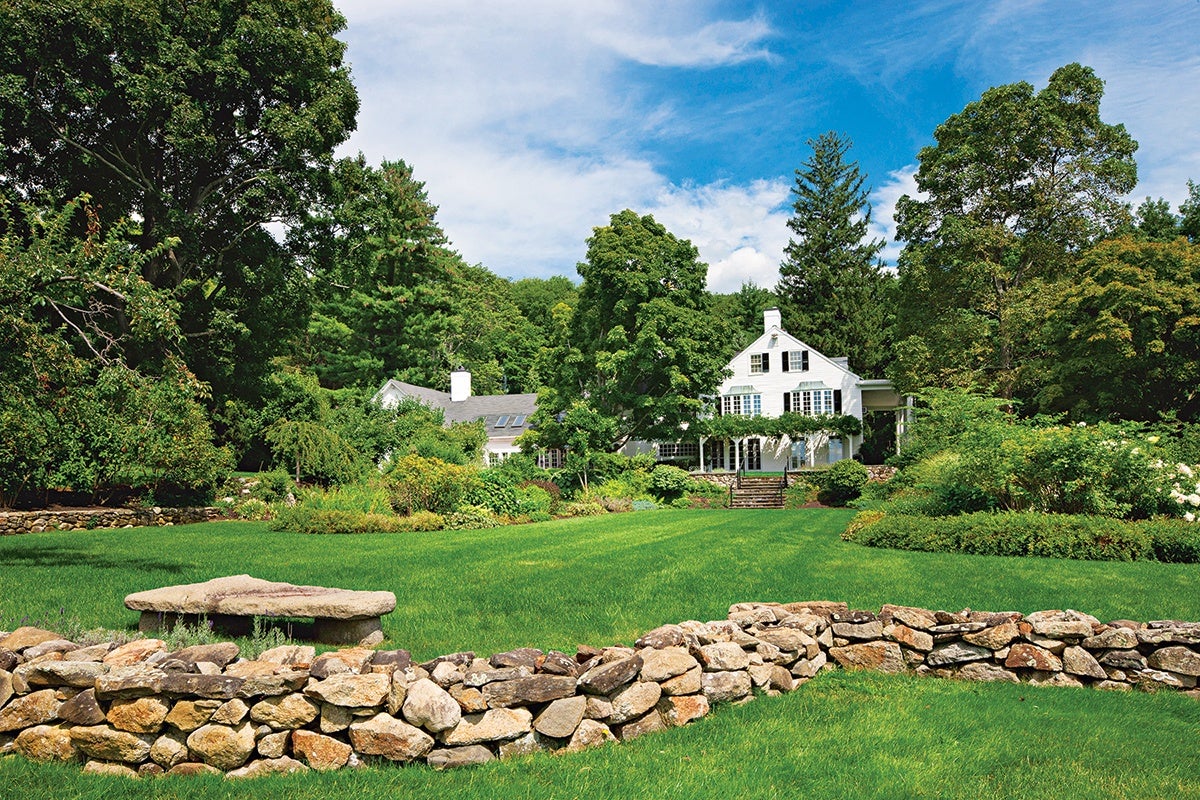 White two-story house with black shutters surrounded by lush green trees and bushes, set behind a large, well-maintained lawn and a low stone wall in the foreground under a partly cloudy blue sky.