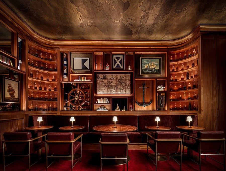 A cozy, nautical-themed room with dark wood paneling and shelves filled with small bottles. The back wall features framed maritime artwork, including a ship's wheel, an anchor, and vintage maps. Five round wooden tables each have a small lamp, surrounded by burgundy upholstered chairs and a matching bench. The ceiling has a textured, aged look, and the floor is covered with a red carpet. The overall lighting is warm and inviting.
