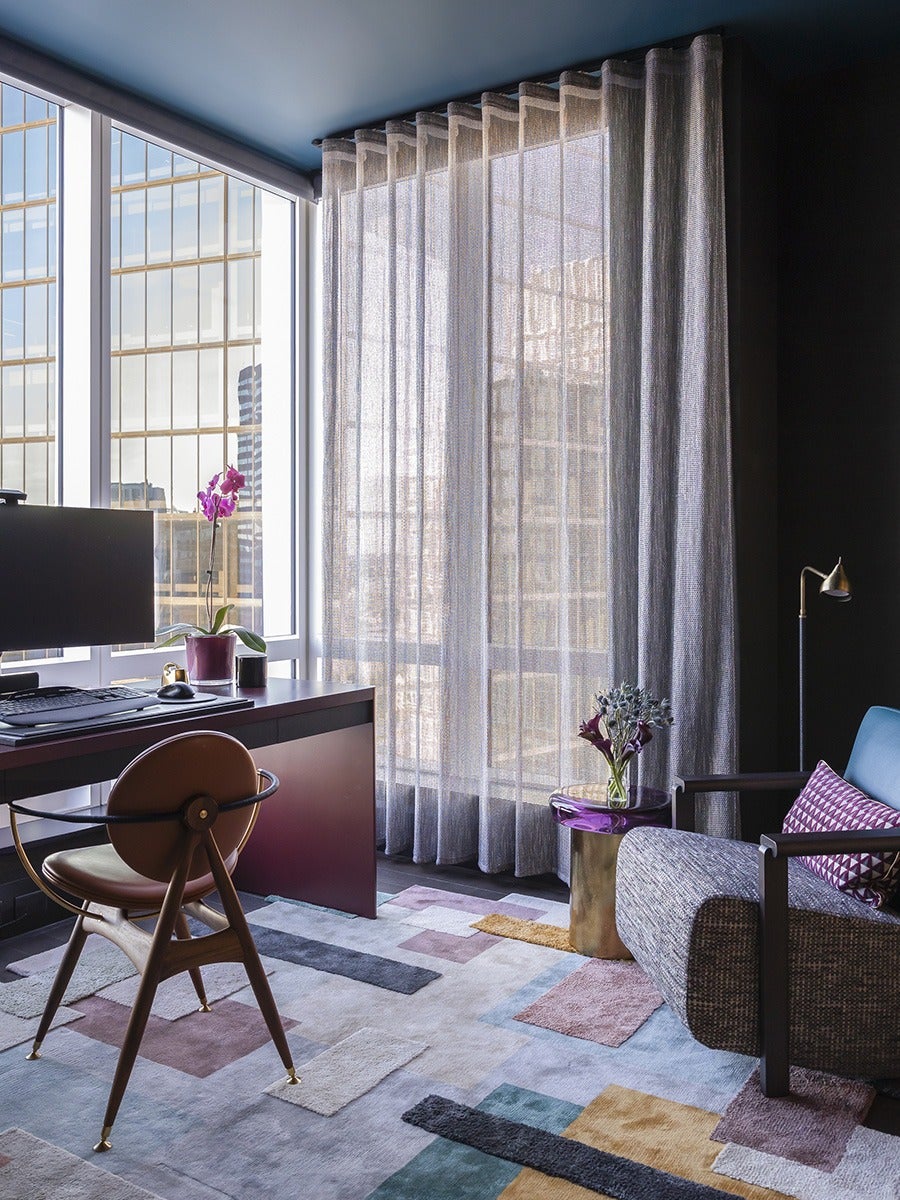 Modern office space with large floor-to-ceiling windows covered by sheer gray curtains. A dark wooden desk holds a computer monitor, keyboard, mouse, and a purple orchid plant. A mid-century style chair with a brown leather seat and wooden legs is positioned at the desk. The floor features a colorful geometric patterned rug. To the right, a textured armchair with a blue cushion and a purple patterned pillow sits next to a small round side table with a purple top and gold base, holding a vase of flowers. A tall, slender floor lamp with a brass finish stands behind the armchair.