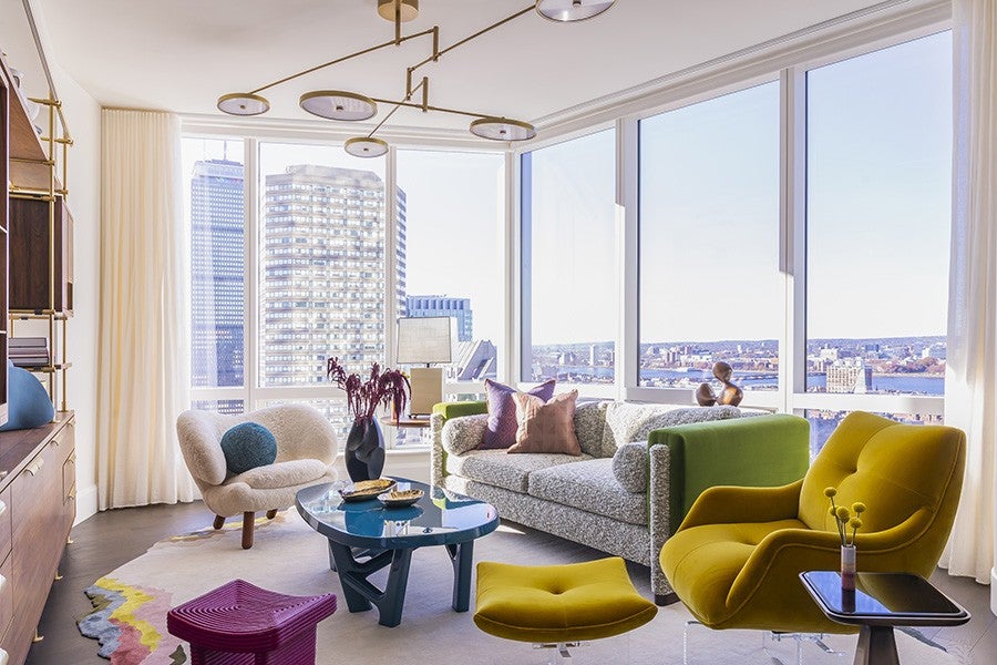 Bright living room with large floor-to-ceiling windows offering a cityscape view. The room features a textured gray sofa with green armrests, a mustard yellow armchair with matching ottoman, a cream-colored rounded chair with a blue cushion, and a blue glass coffee table. A modern chandelier hangs from the ceiling, and a colorful rug with pink, yellow, and gray accents covers the floor. A small side table with a vase holding yellow billy balls is next to the armchair.