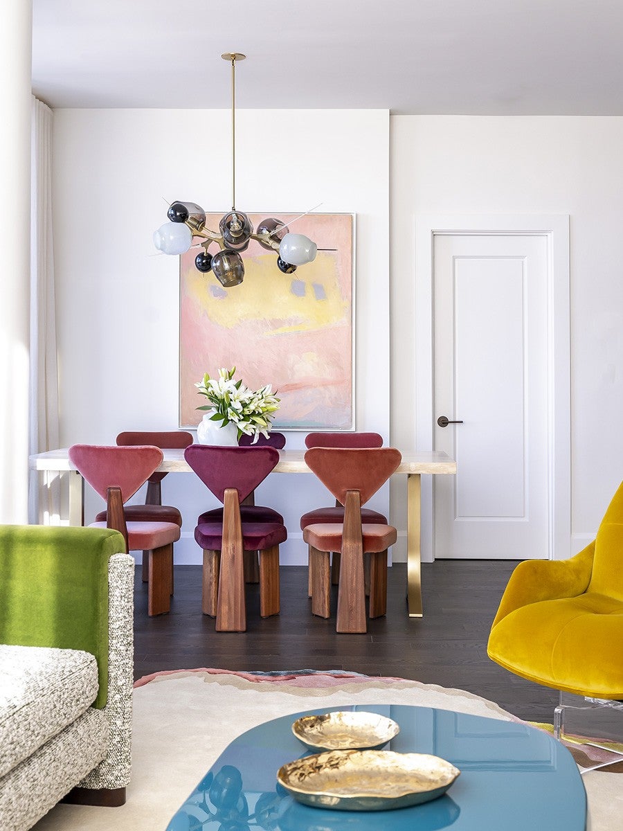 A modern dining area with a rectangular table featuring gold legs and a light-colored top. Six velvet chairs in shades of pink, burgundy, and rust surround the table. A white vase with white flowers sits on the table. Above, a contemporary chandelier with multiple glass globes hangs from the ceiling. Behind the table, a large abstract painting in pastel pink, yellow, and gray tones is mounted on a white wall. The room also includes a green and beige textured sofa on the left, a bright yellow armchair on the right, and a blue glossy coffee table with two gold decorative trays in the foreground. The floor is dark wood, and a light-colored rug with a subtle pink border is partially visible.