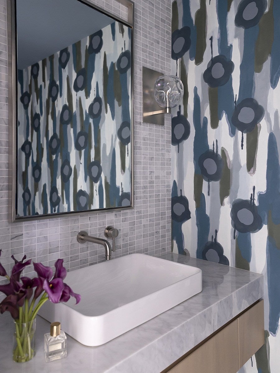 Bathroom countertop with a rectangular white sink, a wall-mounted stainless steel faucet, and a large mirror reflecting a blue and gray floral patterned wall. A small glass vase with purple flowers and a perfume bottle are placed on the marble countertop. A modern wall sconce with a clear glass globe is mounted next to the mirror.
