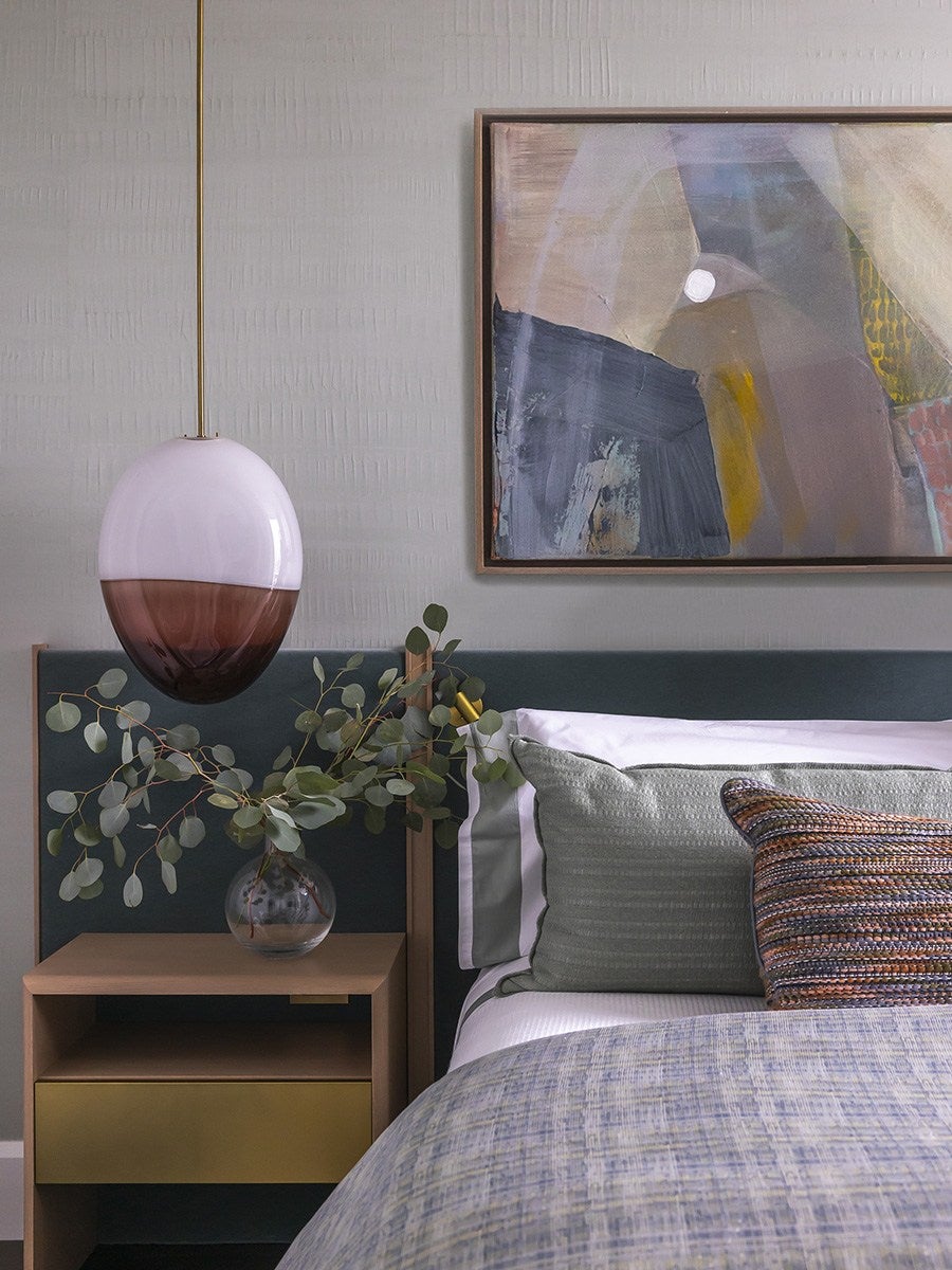 A modern bedroom corner featuring a wooden nightstand with a yellow drawer, a round glass vase holding eucalyptus branches, and a bed with a dark green upholstered headboard. The bed is dressed with white sheets, a light green textured pillow, and a multicolored striped pillow. Above the bed hangs an abstract painting in muted tones of gray, beige, yellow, and blue. A pendant light with a half white, half brown glass shade hangs above the nightstand. The wall behind is painted in a soft gray-green color with subtle texture.