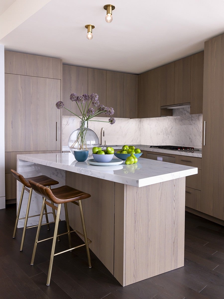 Modern kitchen with light wood cabinetry and a white marble countertop island. Two wooden bar stools with brass legs are positioned at the island. On the island, there is a clear glass vase with purple flowers, a small blue bowl, and two larger bowls filled with green apples and pears. The backsplash is white marble, matching the countertop, and the floor is dark wood. Two brass ceiling light fixtures with exposed bulbs are visible.