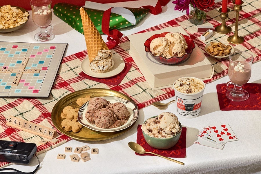 A table set with various desserts and snacks on a red and white checkered tablecloth. There are bowls of ice cream, a plate with chocolate chip cookies and a waffle cone, a cup of hot chocolate with marshmallows, a bowl of popcorn, and a small bowl of crackers. A Scrabble board with tiles spelling "ICE CREAM," "CONE," and "SALT STRAW" is visible, along with scattered Scrabble tiles. Also on the table are playing cards, a camera, two brass candlesticks with red candles, a green party hat with gold hearts, and a small vase with purple flowers.