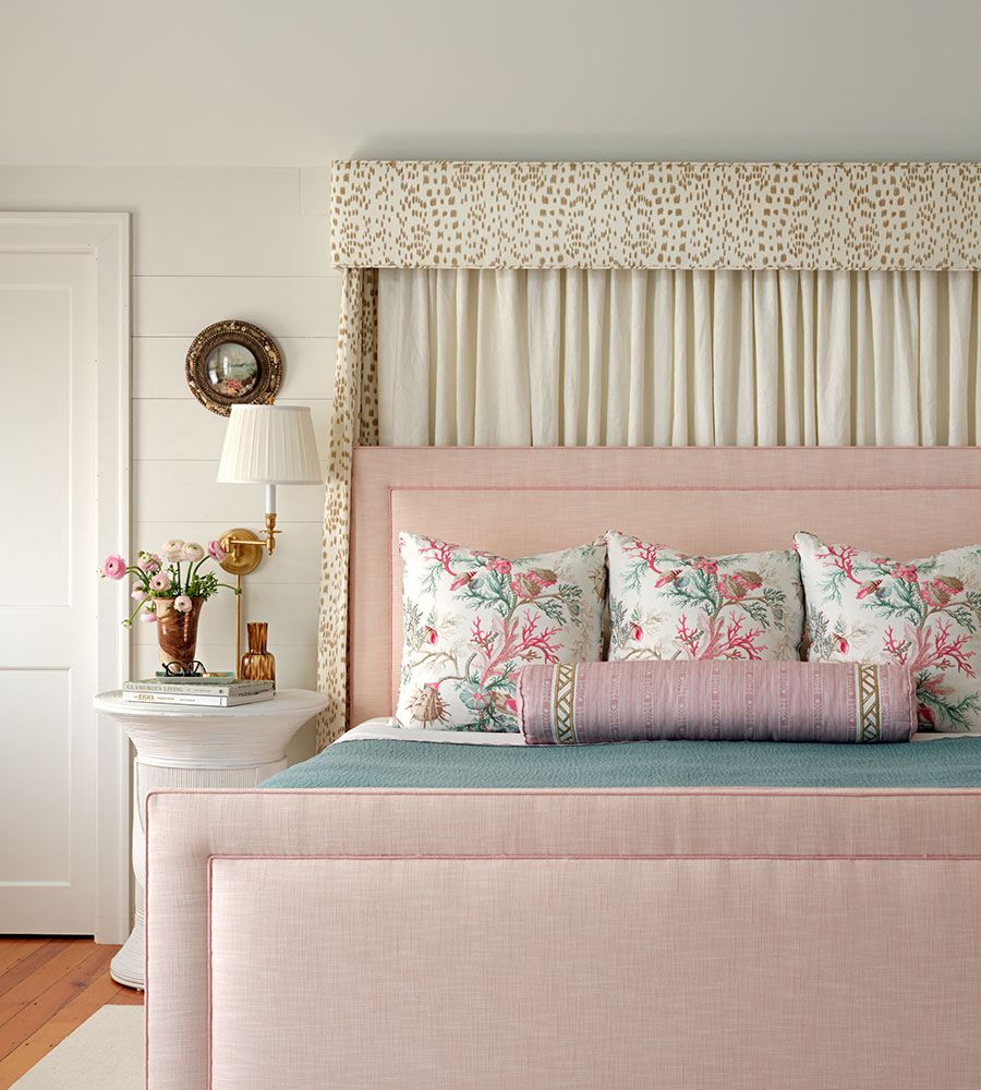 A bedroom features a pink upholstered bed with a tall headboard and footboard. The bed is adorned with three floral-patterned pillows in shades of pink and green, and a long cylindrical bolster pillow in a muted pink tone with decorative trim. Behind the bed is a fabric-covered canopy frame with beige and white patterned fabric and cream-colored curtains. To the left of the bed is a white round side table holding a copper vase with pink and white flowers, a small amber glass vase, and two stacked books. A brass wall-mounted lamp with a white pleated shade is above the table. The room has white shiplap walls and a wooden floor.