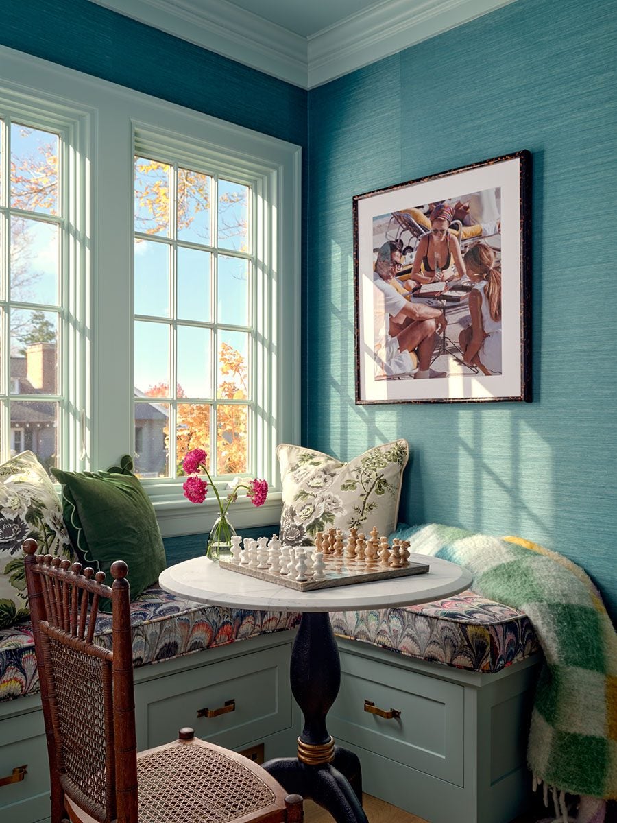 Cozy corner with a round white marble-top table holding a chessboard and a small vase with pink flowers. Surrounding the table is a built-in bench with floral and green cushions, a green and white checkered blanket, and a wooden chair with a woven seat. The walls are teal with a framed photo of people playing a board game. Large windows let in natural light, casting shadows on the wall.