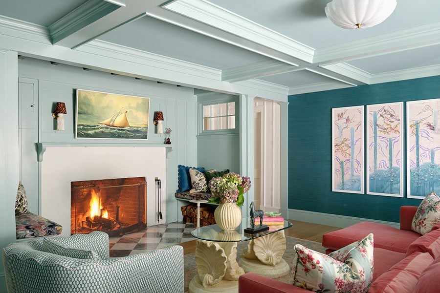 Living room with light blue walls and ceiling beams, featuring a lit fireplace with a sailboat painting above it. The room includes a patterned armchair, a coral sectional sofa with floral pillows, a glass coffee table with a vase of flowers and decorative items, and a blue accent wall adorned with a four-panel artwork depicting birds and branches.