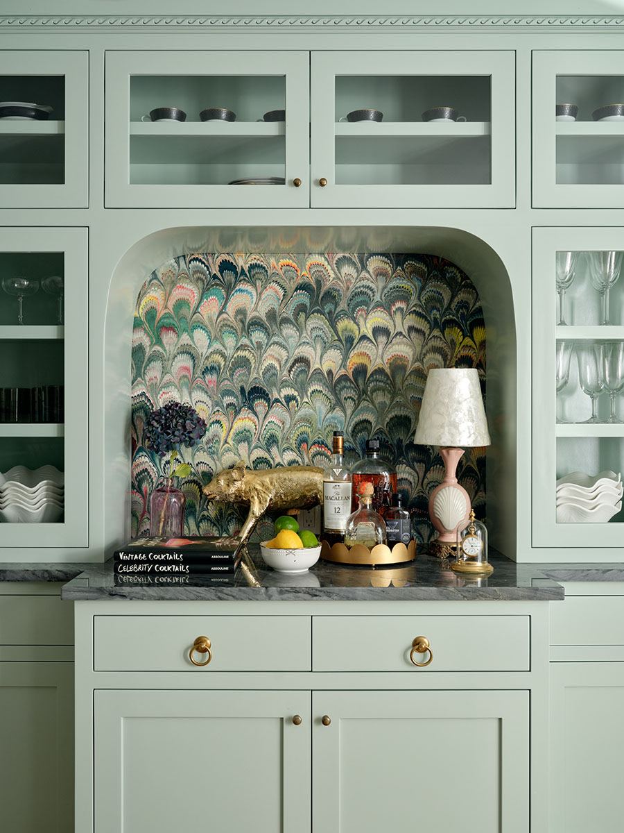 A mint green built-in cabinet with glass doors displaying cups, plates, and glasses. The countertop is dark marble with a decorative alcove featuring a colorful, peacock feather-patterned wallpaper. On the countertop, there is a gold pig sculpture, a bowl with lemons and limes, a tray with various liquor bottles, a pink lamp with a white shade, a small clock under a glass dome, and two stacked books titled "Vintage Cocktails" and "Celebrity Cocktails.