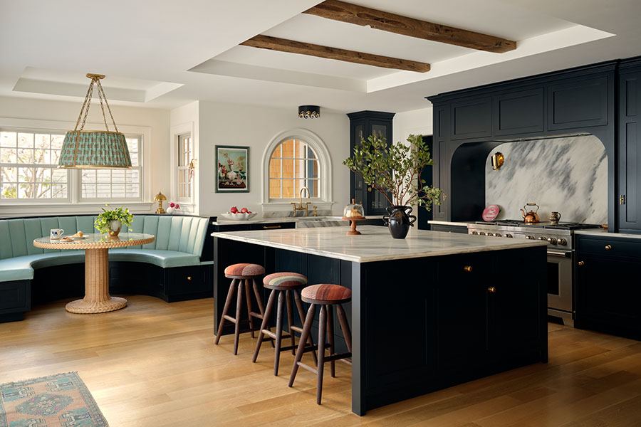 Kitchen with dark navy cabinetry and a large marble island featuring three wooden stools with striped cushions. A curved light blue banquette surrounds a round wicker table under a large woven pendant light. The space has light wood flooring, exposed wooden ceiling beams, and a marble backsplash behind the stove. A black vase with greenery decorates the island.