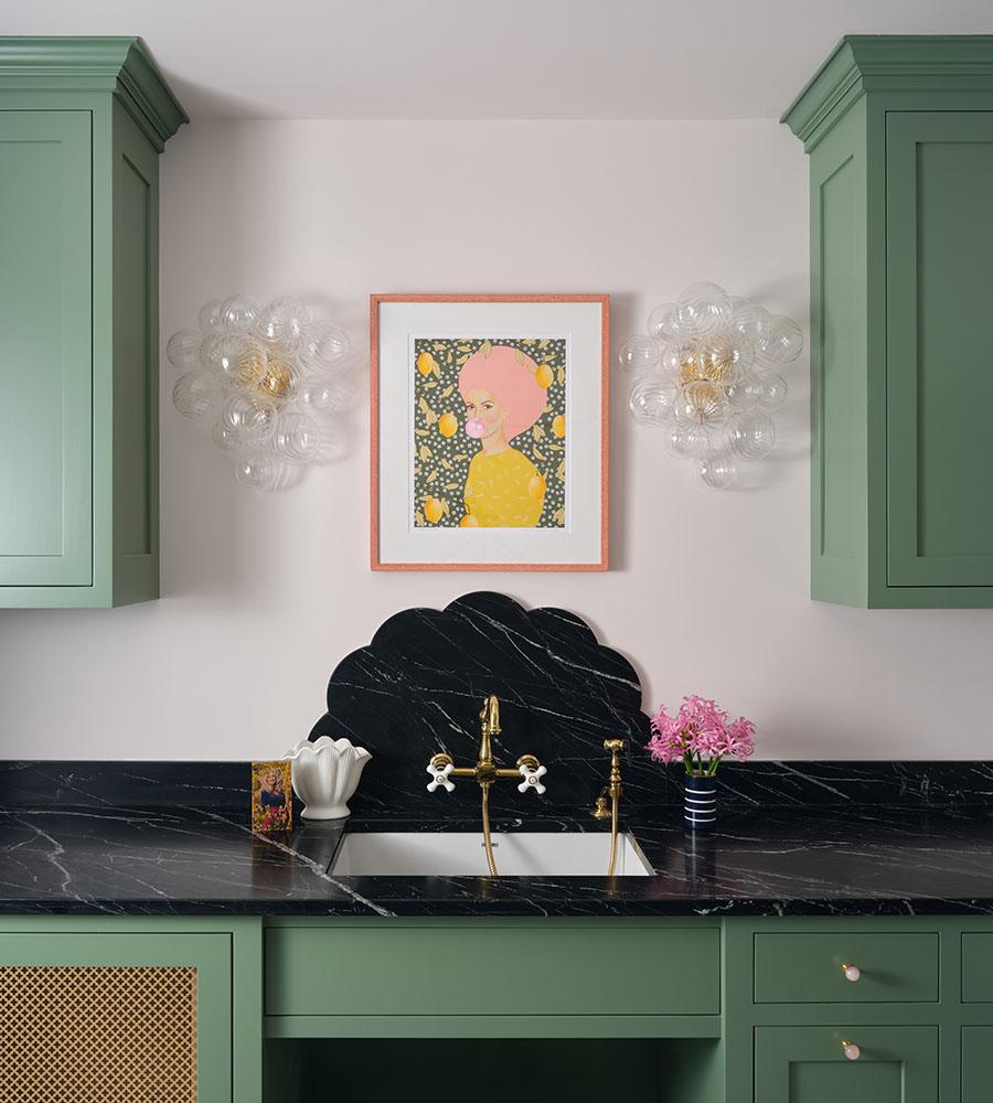 A kitchen sink area with green cabinetry and a black marble countertop and backsplash featuring a scalloped design. Above the sink is a framed artwork of a woman with pink hair blowing a bubblegum bubble, set against a patterned background with yellow lemons. On either side of the artwork are two clear bubble-like wall sconces with gold fixtures. A small white scalloped vase and a small striped vase with pink flowers are placed on the countertop.