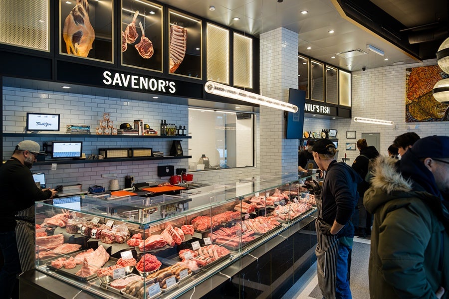 Butcher shop interior with a glass display case filled with various cuts of raw meat. The shop has white subway tile walls and black shelving holding items like books, bottles, and boxes. Above the counter, illuminated signs read "SAVENOR'S" and "WULF'S FISH." Several customers and a butcher wearing a cap and apron are present. The ceiling has modern lighting fixtures, and framed images of meat cuts are displayed above the signs.