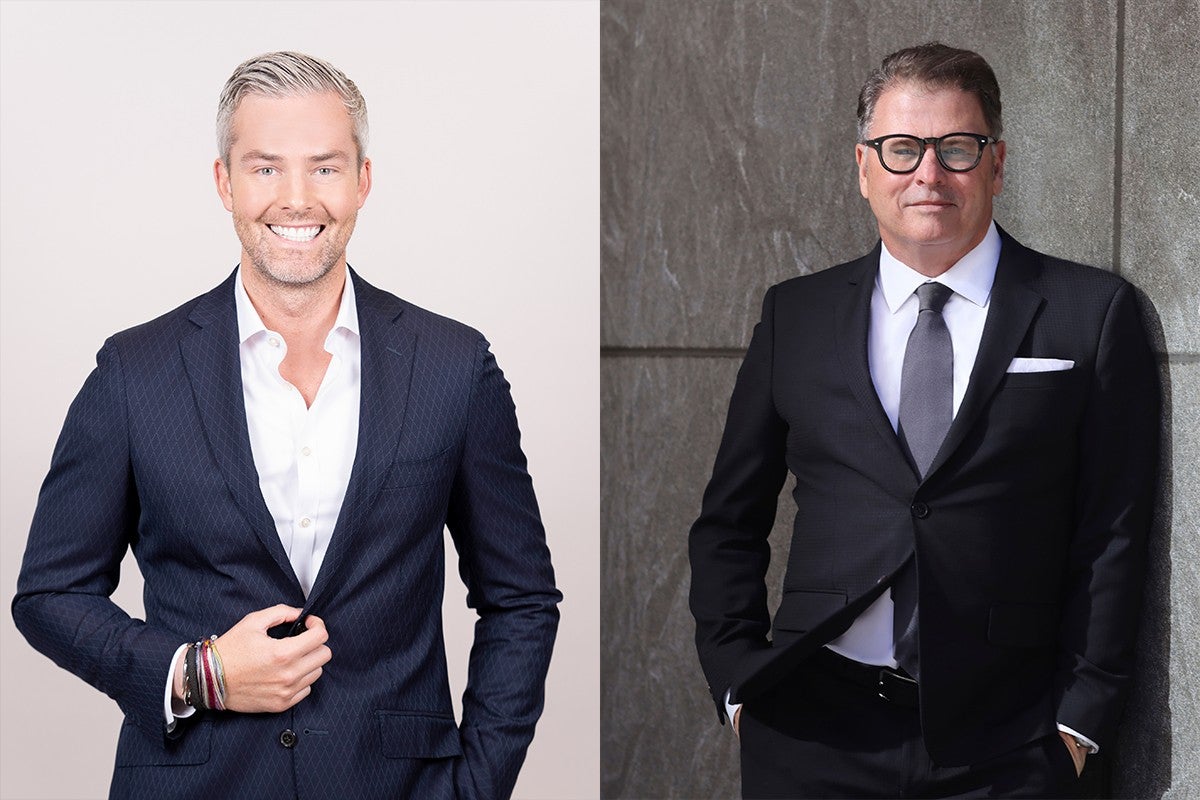 Two men dressed in suits are shown side by side. The man on the left has short, light gray hair and is smiling broadly. He wears a dark patterned suit jacket over a white shirt, with several bracelets on his left wrist. The man on the right has short dark hair, wears glasses, and has a neutral expression. He is dressed in a black suit with a white shirt, gray tie, and a white pocket square, standing against a textured stone wall with his hands in his pockets.