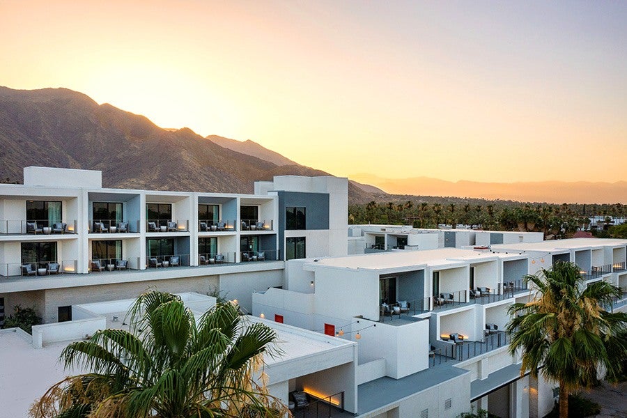 Modern white multi-story hotel building with balconies, set against a backdrop of mountains at sunset, with palm trees in the foreground.