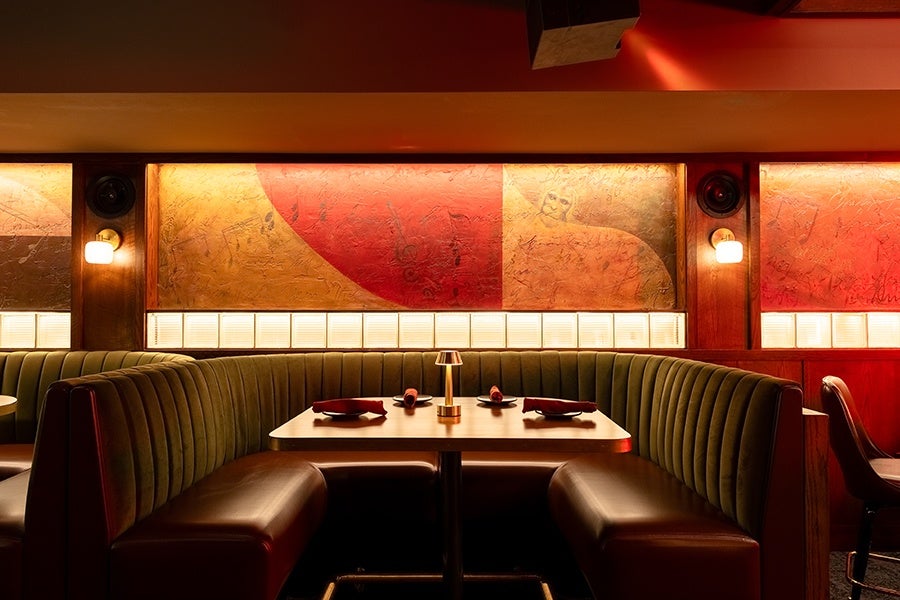 A cozy restaurant booth with dark green upholstered seats and a wooden table set with folded red napkins and small plates. The background features warm, ambient lighting with wall sconces and abstract red and gold artwork on the walls.