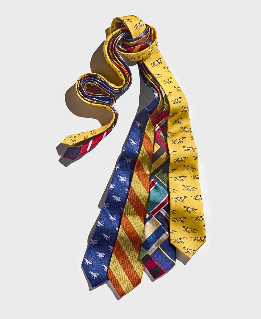 Five silk neckties are displayed, each with a distinct pattern and color scheme. From left to right: a yellow tie with small black horse illustrations, a blue tie with white airplane motifs, a yellow and orange diagonal striped tie, a multicolored geometric patterned tie with shades of green, red, blue, and beige, and another yellow tie with black horse illustrations. The ties are loosely knotted together at the top and spread out at the bottom against a light gray background.