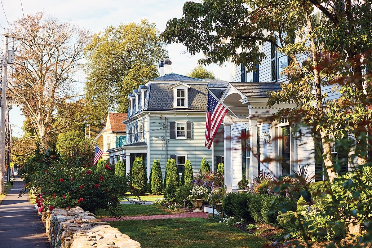 Two traditional American houses with light-colored siding are shown along a tree-lined street. Both houses have American flags displayed near their entrances. The front yards feature well-maintained gardens with shrubs, flowers, and small trees, bordered by a low stone wall and a brick pathway. The scene is bathed in warm sunlight, highlighting the greenery and autumn foliage.