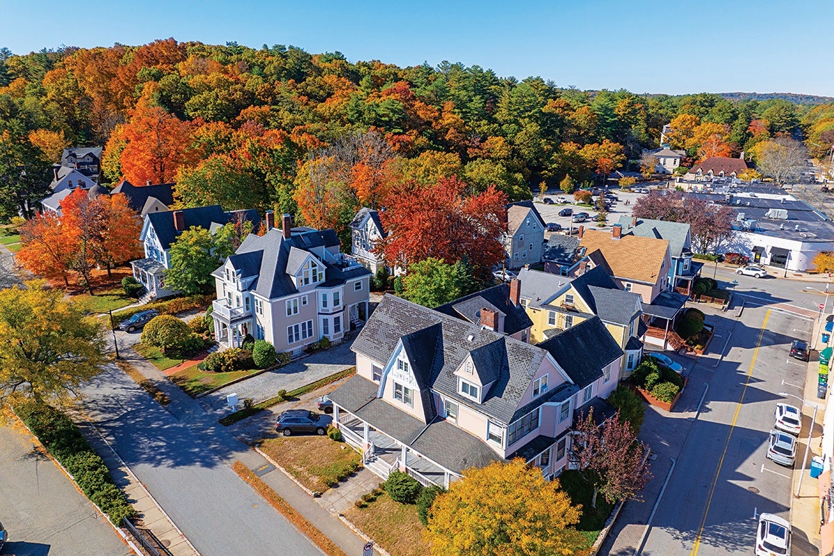 A small neighborhood with several houses featuring pitched roofs and porches, surrounded by trees with vibrant autumn foliage in shades of orange, red, and yellow. The streets are lined with parked cars, and a parking lot with more vehicles is visible in the background. The scene is set under a clear blue sky.