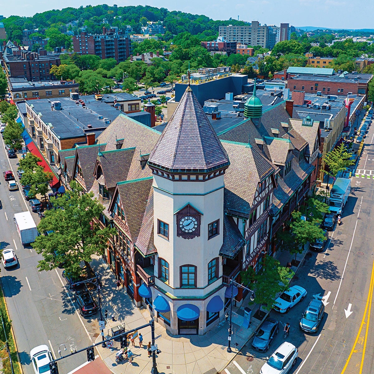 A large, historic building with a steep, multi-gabled roof and a prominent clock tower at the corner. The clock tower has a pointed, shingled roof and four clock faces. The building features Tudor-style architecture with timber framing and cream-colored walls. Blue awnings cover the ground-floor windows and entrances. The building is situated at a busy urban intersection with cars, pedestrians, and trees lining the streets. Surrounding the building are other urban structures and greenery in the background.