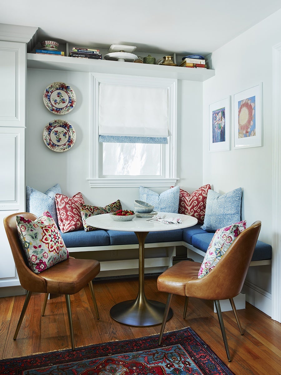 A cozy breakfast nook features a round white marble table with a brass pedestal base, surrounded by a built-in blue cushioned bench and two brown leather chairs. The bench is adorned with a mix of blue, red, and patterned throw pillows. The nook is set against white walls with a window covered by a white Roman shade with a blue trim. Above the window, a shelf holds books and decorative items. Two decorative plates hang on the left wall, and two framed floral artworks are on the right wall. The floor is wooden with a colorful patterned rug partially visible.