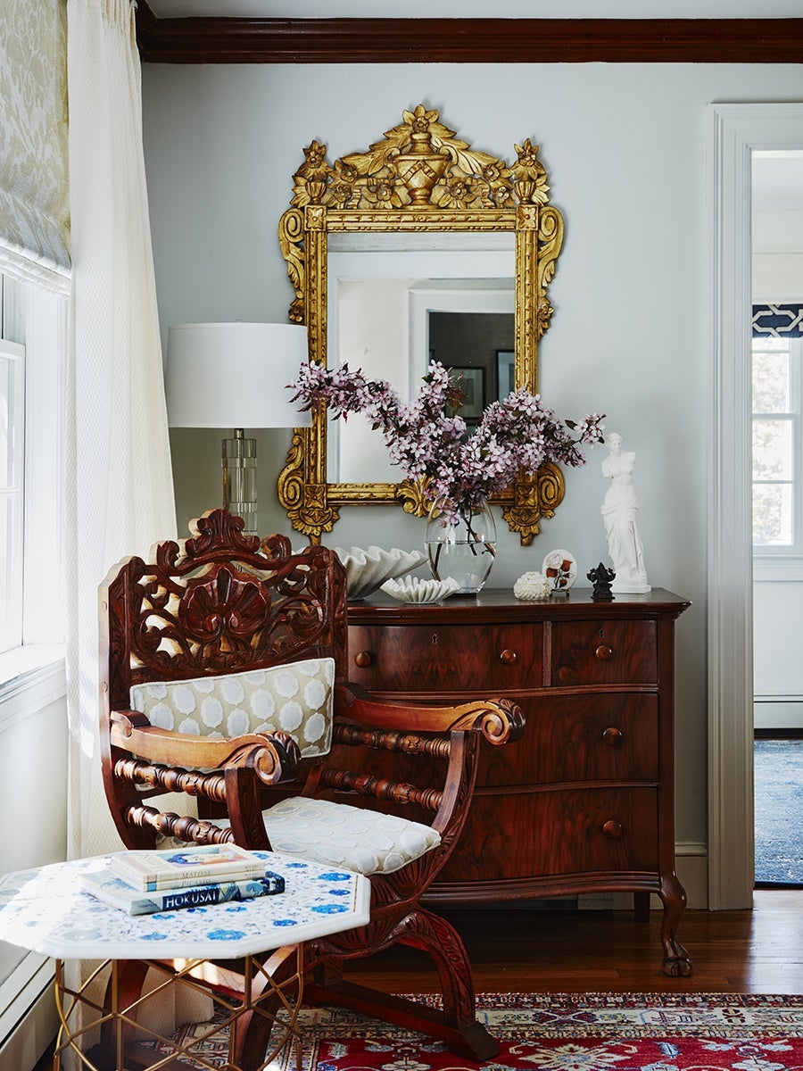 Ornate wooden armchair with light patterned cushions sits next to a small white and blue hexagonal table holding two books. Behind the chair is a dark wooden dresser with a clear vase of pink flowers, decorative shells, and small sculptures. Above the dresser hangs a large, intricately carved gold-framed mirror. The room features light walls, a window with white curtains, and a red patterned rug on a wooden floor.
