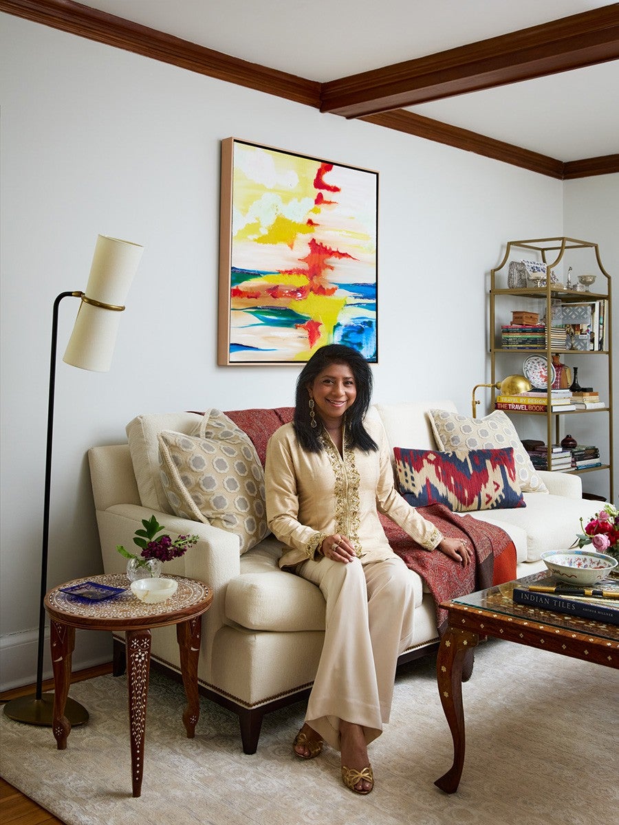 A woman dressed in a beige traditional outfit sits on a cream-colored sofa adorned with patterned and colorful cushions and a red throw. The room features a wooden side table with floral inlay, a tall floor lamp with a white shade, and a wooden coffee table with books and a decorative bowl. Behind the sofa, a vibrant abstract painting with yellow, red, and blue hues hangs on the white wall. A gold metal shelving unit filled with books and decorative items stands in the corner. The room has wooden crown molding and a light-colored rug.