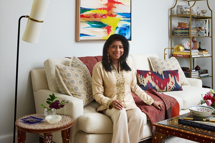 A woman with dark hair wearing a beige embroidered top and light-colored pants is sitting on a cream-colored sofa. The sofa is decorated with patterned cushions and a red throw. Behind her, there is a colorful abstract painting on the wall. To the left, a tall floor lamp stands next to a small round wooden table with a glass vase holding purple flowers and a small bowl. On the right, a glass-top wooden coffee table holds books and a decorative bowl, and a gold metal bookshelf filled with books and decorative items is visible in the background.