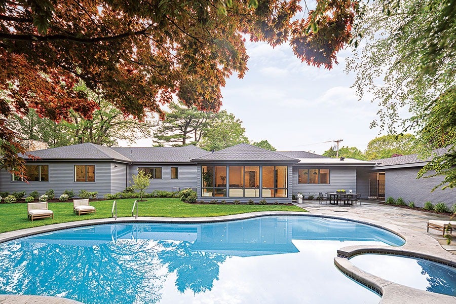 A modern single-story house with gray siding and large windows overlooks a curved swimming pool and a hot tub. The backyard features a well-maintained lawn with two lounge chairs on the grass and a patio area with a dining table and chairs. Trees with green and reddish leaves frame the scene.