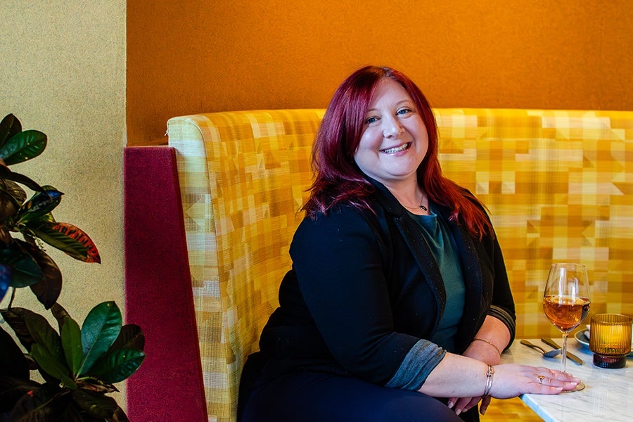 A woman with red hair wearing a black blazer and teal top is sitting on a yellow patterned booth seat. She is smiling and resting her arm on a white marble table with a glass of rosé wine, a yellow candle holder, and cutlery. There is a plant with green leaves on the left side of the image. The background features a warm orange wall.
