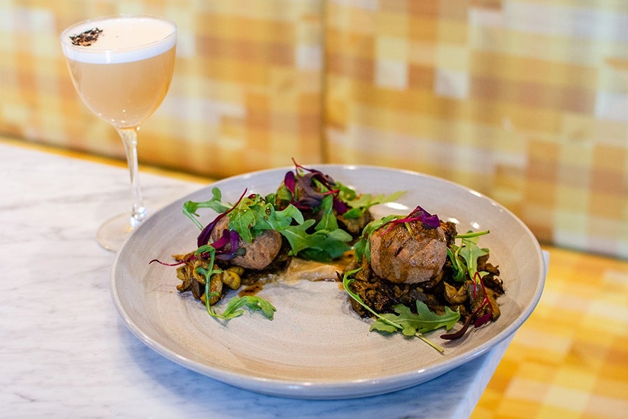 Two small portions of a gourmet dish featuring cassava dumplings on a bed of sautéed mushrooms and greens, garnished with fresh arugula and purple microgreens, served on a beige ceramic plate. Next to the plate is a cocktail in a stemmed glass with a frothy top and a small garnish. The setting includes a marble table and a yellow checkered background.