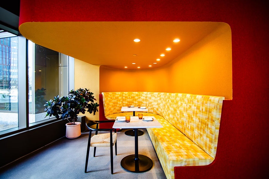 A cozy dining nook with a curved yellow and white checkered bench against an orange wall. Two small square tables with black pedestal bases are set with plates, napkins, and amber-colored glasses. A single black chair with a woven seat is positioned at the nearest table. The space is illuminated by recessed ceiling lights and has a large window on the left side with a potted plant nearby. The surrounding wall is deep red, creating a warm and inviting atmosphere.