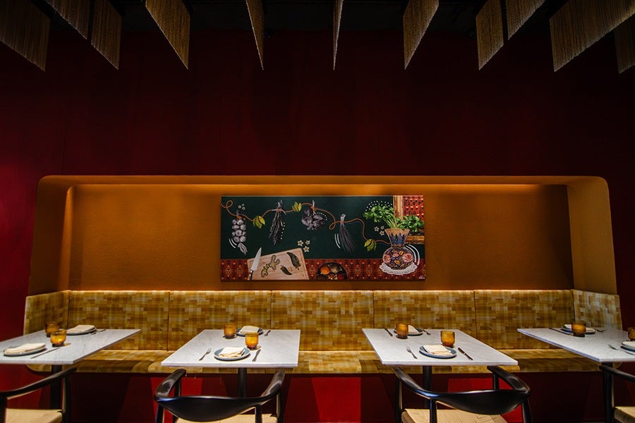 A dining area with a long mustard-yellow cushioned bench against a deep red wall. Four white marble tables are set with plates, napkins, forks, knives, and amber-colored glasses. Above the bench hangs a colorful painting featuring botanical and kitchen-themed elements. The ceiling has hanging fringed decorations. Black chairs with light-colored seats are placed at the tables.