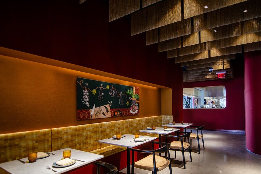 Modern restaurant interior with a row of small square tables set with plates, napkins, and amber-colored glasses. The seating includes a long cushioned bench with a yellow and brown checkered pattern and black chairs with woven seats. The walls are painted in deep red and orange tones, featuring a colorful painting of vegetables and kitchen items. The ceiling has layered, textured hanging panels with recessed lighting. A window offers a view into the kitchen area.