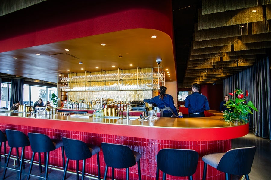 A modern bar with a curved red counter and black bar stools. Behind the counter, two bartenders in dark blue shirts are working, with shelves of glassware and bottles illuminated by warm lighting. The ceiling features layered, textured panels, and there is a large green plant with red flowers on the right side of the bar. In the background, some patrons are seated near large windows.