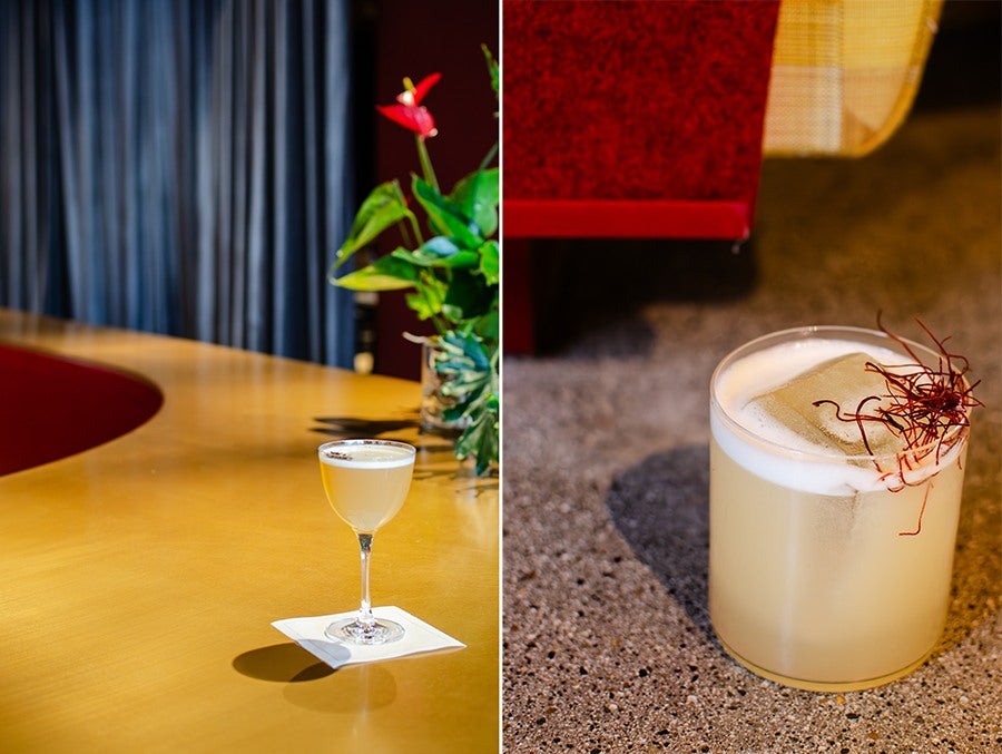 Left side: A cocktail in a coupe glass with a frothy top, placed on a white napkin on a smooth wooden bar counter, with a blurred background featuring a red flower and green leaves. Right side: A pale yellow cocktail in a short glass with a large ice cube, topped with a frothy layer and garnished with thin red strands, set on a speckled countertop with a red and yellow background.